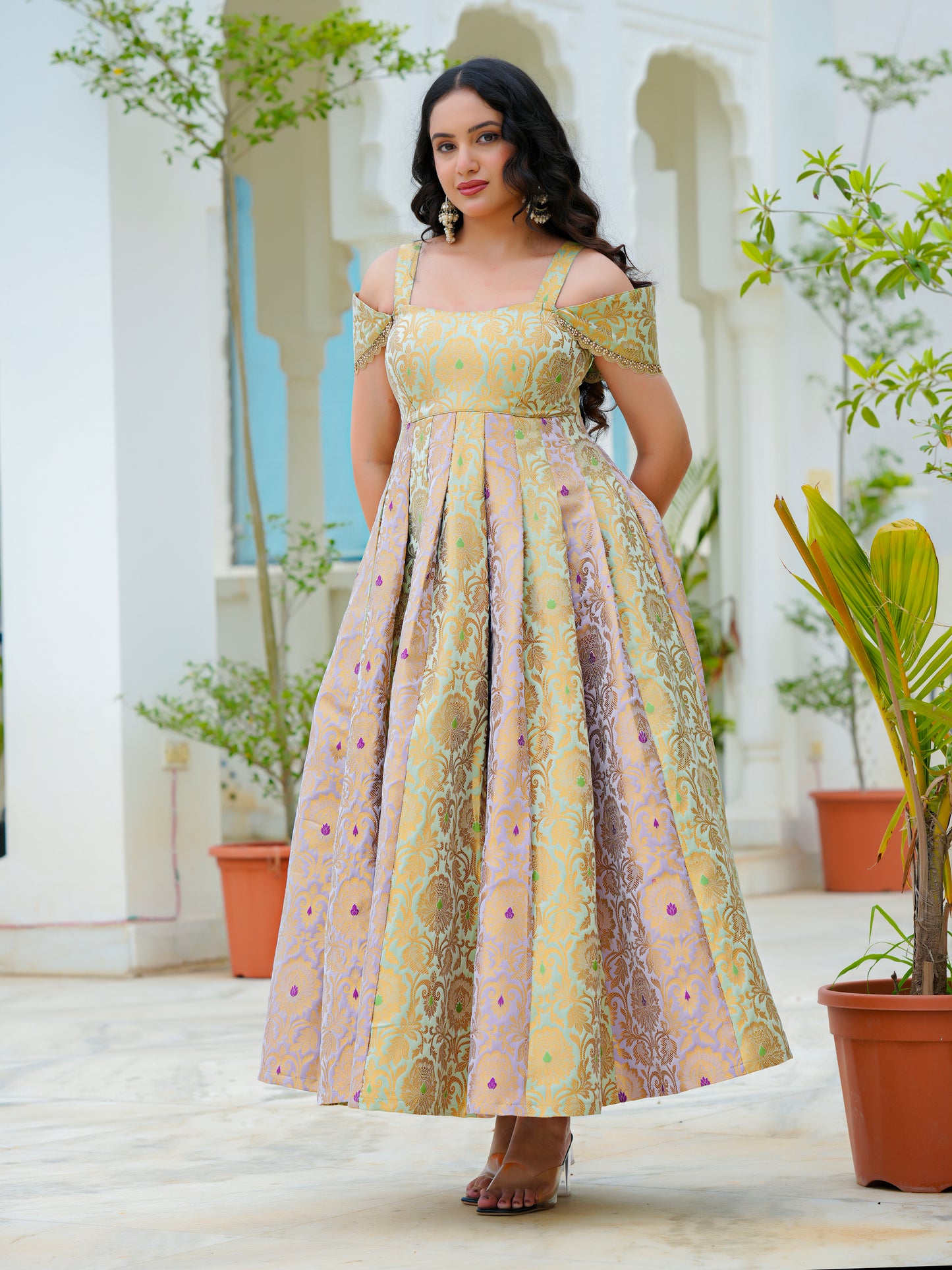 A woman modeling a long off-the-shoulder dress with a yellow, gold, and pink floral pattern, standing in a courtyard with white arches and potted plants.