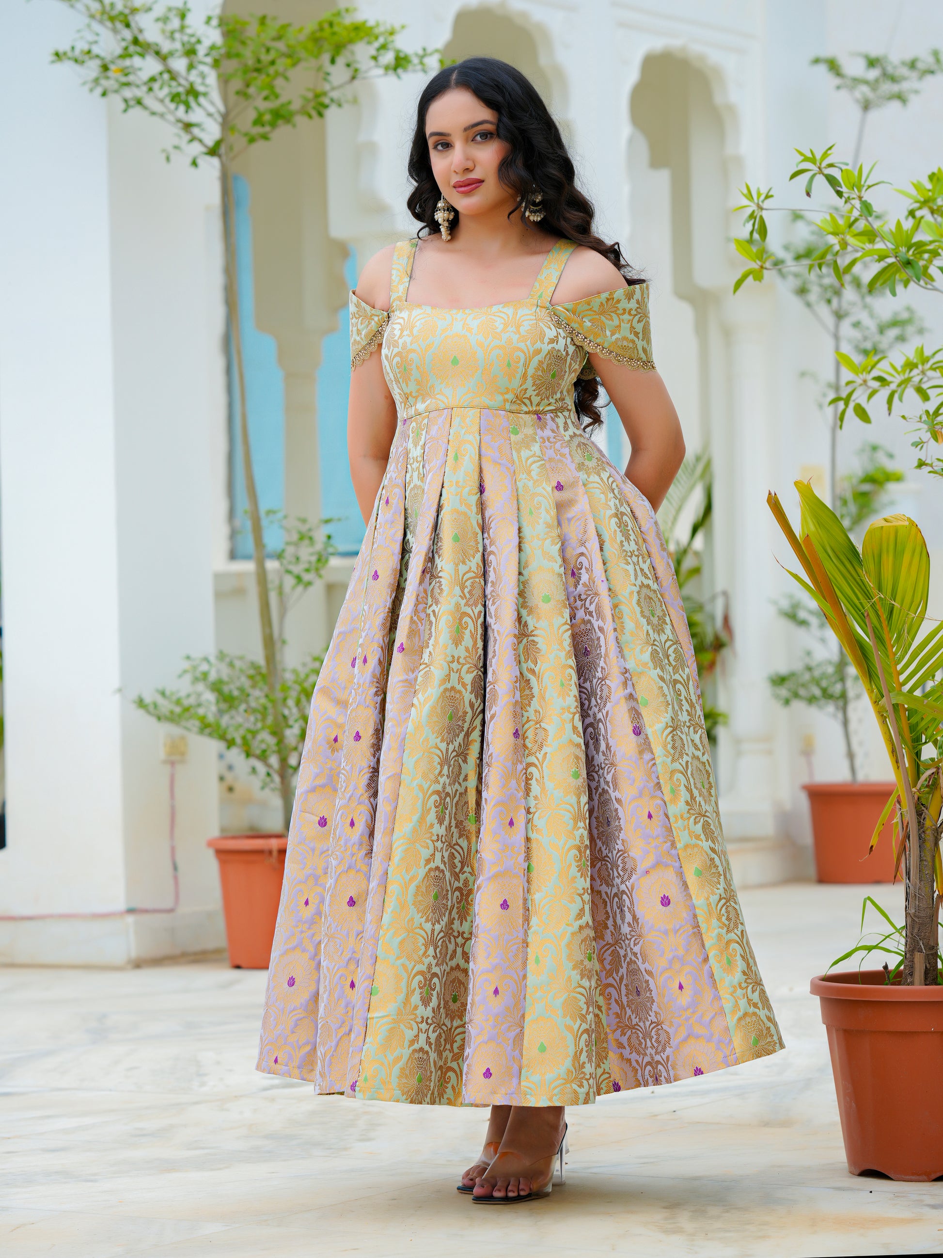 A woman modeling a long off-the-shoulder dress with a yellow, gold, and pink floral pattern, standing in a courtyard with white arches and potted plants.