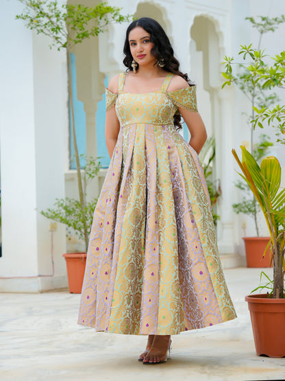 A woman modeling a long off-the-shoulder dress with a yellow, gold, and pink floral pattern, standing in a courtyard with white arches and potted plants.